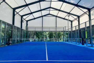 Blue padel court enclosed by glass and fencing beneath a translucent roof